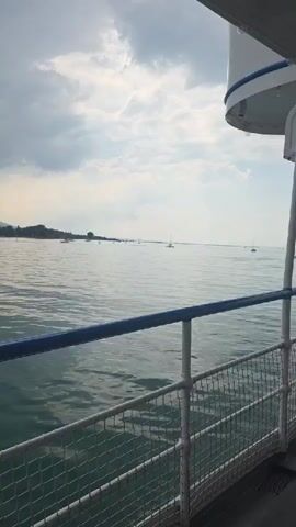 The video shows a view from a boat on a large body of water. The water is calm with some gentle ripples. The sky is overcast with dramatic clouds, suggesting potential for rain or stormy weather, although the light is still bright. In the distance, a shoreline with trees is visible, and several small boats are scattered across the water. The perspective is from the deck of the boat, looking out over the water and towards the distant land. A railing with blue and white accents is in the foreground, partially obscuring the view. The overall impression is one of a slightly moody but scenic boat journey.