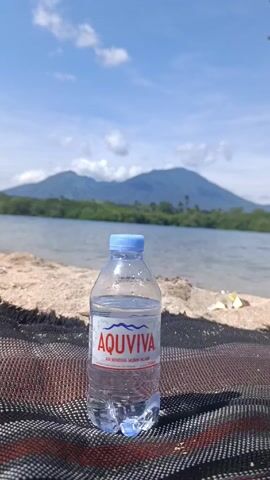 A small plastic bottle of AQUVIVA mineral water rests on a brown and black woven surface. Behind it, there is a beach with sand, and beyond the beach, water stretches to a mountainous island in the background. The sky is blue with scattered white clouds.