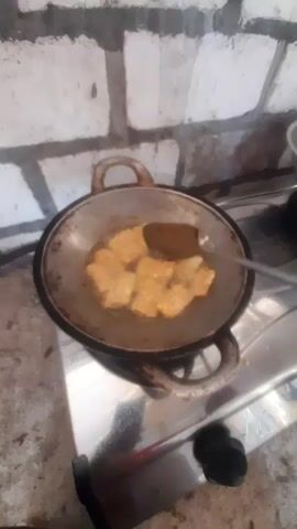 A hand holds a metal spatula, flipping pieces of golden-brown food in a metal frying pan. The pan sits on a gas stovetop, with a flame visible underneath. The background shows a brick wall. The food appears to be fried, possibly tempeh or tofu. The lighting is dim, and the image quality is low.