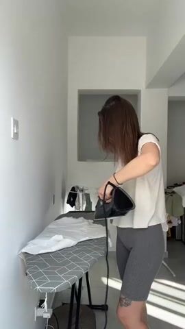 A young woman with shoulder-length brown hair is ironing a white garment on a black and grey ironing board. She is wearing a light-colored t-shirt and grey ribbed shorts. The ironing board is set up against a white wall with a light switch visible. Behind the ironing board, a small gray recessed shelf is visible. Sunlight streams in from the right, casting shadows and illuminating the scene. The woman appears focused on her task. In the background, a laundry basket and other clothes are visible, suggesting a domestic setting. The overall impression is one of everyday chores being performed in a bright, modern interior. There is no visible text in the media file. The context provided suggests the location is Limassol, Cyprus. The lighting indicates it is daytime.