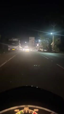 A first-person perspective video shows a person riding a scooter at night on a road. The road is lit by streetlights and the headlights of passing vehicles. There are several buses, cars, and other vehicles on the road. The rider is approaching a busy intersection with traffic signals. The video is dark and grainy due to the low light conditions.