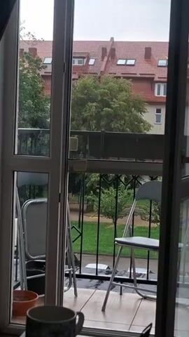 A view through a glass door of a balcony with two chairs and a potted plant. Beyond the balcony, a green lawn and trees are visible, with buildings in the background. The sky is overcast.