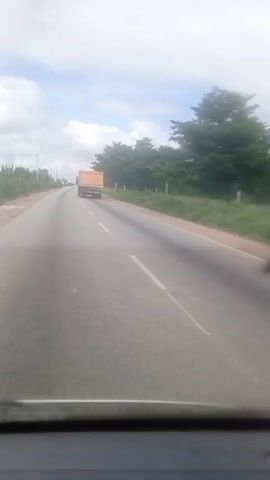 The video shows a view from inside a vehicle moving on a road. An orange dump truck is driving ahead in the same lane. The road has a white line marking the center and is flanked by green vegetation and trees. The sky is partially cloudy. The video appears to be shot during daylight.