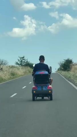 A person is riding a blue and black mobility scooter down a paved road. The person is facing away from the camera and is wearing a dark shirt and a baseball cap. The mobility scooter has a red decal on the backrest and has turn signals on. The road is flanked by dry grass and some scattered trees. The sky is blue with white clouds. The road has a dashed white line dividing the lanes and a solid white line on the right side. A speed limit sign is visible in the distance on the right side of the road. The video is filmed from a slightly elevated position, possibly from another vehicle.