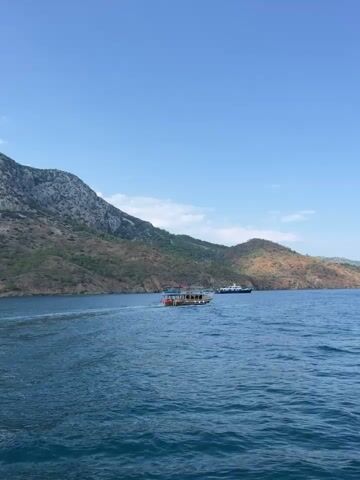 The video shows a blue sky and calm blue water. In the foreground, there is a boat with people on it. In the background, there are mountains and other boats. The scene appears to be a sunny day in a coastal area.