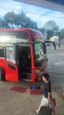 A red bus is parked at a gas station. The bus doors are open, and several people are near it. One person is carrying two bags, walking away from the bus. Another person is looking at the bus's interior. Another person is interacting with luggage located near the bus. The environment appears to be a bright, sunny day with trees and buildings visible in the background.