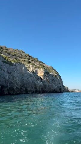 The video captures a boat's perspective as it moves through clear turquoise water near a rocky coastline. A steep, vegetated cliff dominates the left side of the frame, with a man-made structure, possibly a tunnel entrance, carved into its face. The sky is a vibrant, cloudless blue, suggesting a bright, sunny day. In the distance, a hazy shoreline with buildings and masts of sailboats hints at a coastal town or harbor. The water is choppy, indicating the boat is in motion, creating white foam and ripples. The overall scene conveys a sense of exploration and the beauty of a coastal landscape on a pleasant day. No visible text is present.