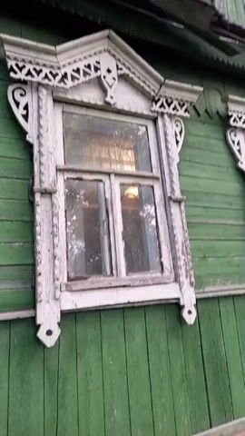 The camera pans upward along the side of a green wooden house. The house has two windows with ornate white frames. Above the windows, the roof line is visible with some damage to the wooden shingles. Above the roof line, the side of the attic has a single small window, and a handle is affixed to the siding. In the background, trees and foliage are visible.
