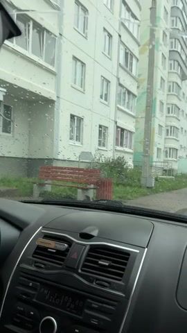The video shows the interior of a car looking out through a rain-streaked windshield at a residential building. In the foreground, the car's dashboard is visible, including the air conditioning vents and a radio display. Outside the car, a red bench and a trash can are visible in front of the building. The building itself is a multi-story apartment complex with numerous windows. The rain is evident on the windshield, creating a blurred effect on the scene outside. The overall impression is of a wet, overcast day.