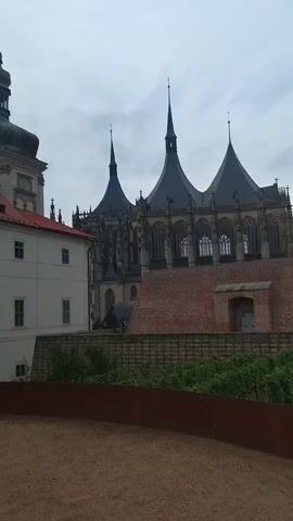 The video opens showing the exterior of a historical building with a red tiled roof and a tall, dark green steeple. In the foreground, a large, brown electrical plug and cord lie on the ground. The camera pans to the left, revealing more of the building's facade and the large plug sculpture. The sky is overcast, and the ground appears to be sandy or dirt. The video continues to show the building and the sculpture from different angles.