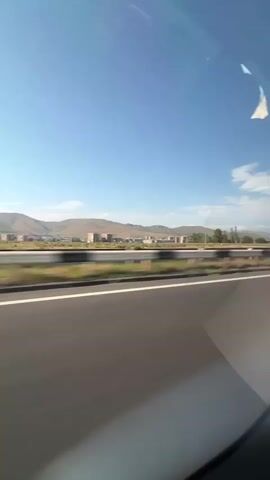 This video shows a view from inside a vehicle, looking out at a highway. The road has a white line marking the lane. In the distance, there are hills and some buildings. The sky is blue with a few clouds. The movement of the camera suggests the vehicle is in motion. There are reflections on the window, including what appears to be a sticker or piece of paper.
