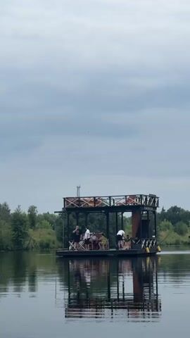 A floating platform with a two-story structure on top is on a calm body of water. The structure is made of dark wood and has open sides with railings. On the lower level, several people are visible standing and sitting around tables. The upper level also appears to have seating. The platform is reflected in the water, which is disturbed by a slight ripple. The background shows a line of trees and a cloudy sky. The overall scene suggests a recreational or dining area on the water.