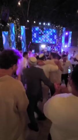 A group of men are dancing in a circle in an outdoor setting at night. They are wearing mostly white clothing, and some are wearing kippahs. There is a large screen in the background displaying a pattern of blue circles. String lights are visible overhead. The men appear to be celebrating, with some hugging and raising their arms. The lighting is dim and colorful, with blue and white lights illuminating the scene.