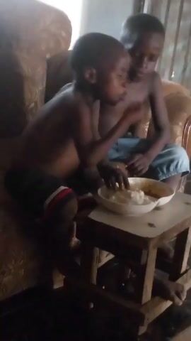 Two young, shirtless boys sit close to each other on a floral-patterned couch. In front of them, on a small, worn, wooden table, is a white bowl containing a white, starchy food alongside a reddish-brown sauce. The boy on the left, wearing black shorts with white and red stripes, picks up a piece of the food and eats it. The boy on the right, wearing light blue shorts and a necklace, is also eating from the bowl. The room seems dimly lit.