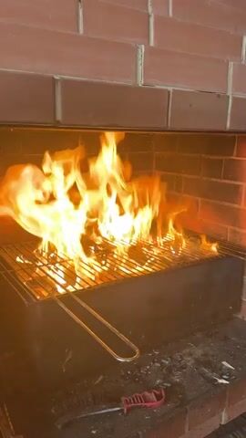 A barbecue grill is positioned inside a brick enclosure. Flames are actively burning on the grill. A metal barbecue tong is resting on the grill. A red-handled barbecue brush is on the ledge below the grill. The enclosure is made of red bricks.