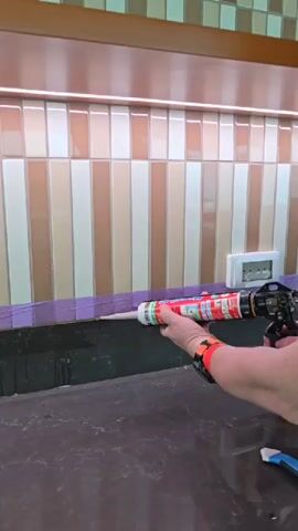 A person is applying sealant with a caulking gun along the edge of a kitchen countertop. The countertop is a dark, speckled material. Above it is a backsplash made of vertical tiles in alternating shades of white and beige. A strip of purple painter’s tape runs along the top edge of the countertop, just below the backsplash. The person is wearing a watch with an orange band on their left wrist. An electrical outlet is visible on the backsplash. The caulking gun is loaded with a tube of sealant that has a red, green, and white label. The lighting in the kitchen is bright, and there is a light fixture visible above the tiles. A blue tool is lying on the countertop.
