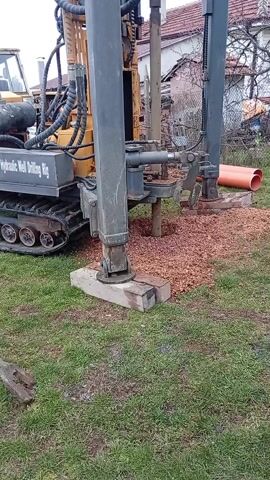 A yellow hydraulic well drilling rig is on a green lawn covered with red gravel. It has a grey base with black treads and is supported by grey legs that are resting on concrete slabs. Behind the rig are tools, including a shovel and metal poles. Beyond the lawn, there is a fence and houses with red tile roofs. The sky is overcast.