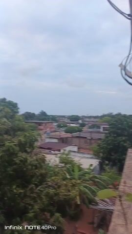 The video shows a rooftop view of a neighborhood at sunset. The sky is cloudy with a pinkish hue around the setting sun. In the foreground, there's an unfinished concrete structure. The middle ground shows residential buildings with various colored roofs and trees scattered throughout the area. A car drives down a street. In the background, there's a communication tower rising above the buildings and trees. The video is shot with an Infinix NOTE 40Pro.