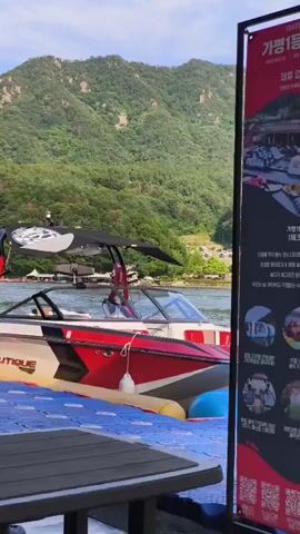 A low-angle medium shot shows a wakeboard boat docked on a blue floating platform. The boat is red, white, and yellow, with "NAUTIQUE" written on its side. A person is partially visible inside the boat's cabin. Behind the boat, a calm lake reflects the sky and the surrounding green, tree-covered mountains. To the right of the frame, a tall red banner displays Korean text and images, likely advertising water sports activities. The sky is partly cloudy.