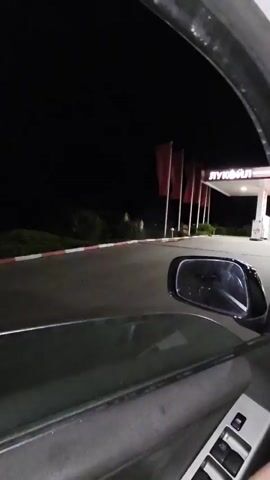 The video shows a person sitting in a car at night, looking out toward a Lukoil gas station. The driver's left arm is visible, wearing a watch with a silver band. The car's interior is dark, and the gas station lights illuminate the area outside. A silver car is parked at one of the pumps. The gas station has a red-and-white color scheme, and several red flags are visible.