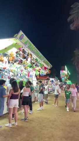 The video captures a lively scene at a fairground at night. The primary focus is on a carnival game stall filled with an abundance of plush toys hanging from the ceiling and displayed on shelves. The stall has a sign overhead that reads "ATRACCIONS DE TIR AMB...(the rest is cut off)". People of various ages are milling around the stall, some looking at the prizes, others appearing to be waiting in line or having just played. The atmosphere is festive, suggested by the bright lighting of the stall and the gathering of people. The time of day is clearly night, with the dark sky visible above the stall's illuminated roof. The ground appears to be dirt or sand, typical of an outdoor fairground. The people are dressed casually in summer attire, indicating warm weather. A young boy in a green "Sky" branded t-shirt is visible on the right side of the frame, adding to the family-friendly feel of the event. The overall mood seems to be one of enjoyment and engagement with the fair activities.
