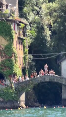 A group of people are gathered on a stone bridge over a body of water. The bridge is arched and covered in ivy. A man in red swim trunks jumps from the bridge into the water, creating a large splash. Several other people are visible on the bridge, some sitting, others standing, and some appear to be watching the jumper. In the background, a building covered in ivy is attached to the side of a hill. There are trees behind the building. The water is a clear blue-green color, and some yellow buoys are visible in the water near the bridge. Other people are in the water below the bridge, swimming. The sun is shining, and it appears to be a warm day.