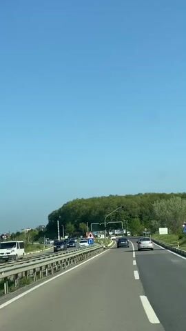 The video shows a roadside scene in sunny conditions, likely captured from a moving vehicle. Several cars are on the road in both directions. A U-turn sign is visible along with traffic lights showing orange. On the right side, a hill is covered with green trees. The sky is clear and blue.