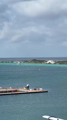 A seaplane with orange and white markings is seen on the water, creating a wake as it moves towards the right of the frame. In the foreground, a dock with railings and several large cylindrical tanks is visible. The water is a deep blue-green, and a distant landmass with some industrial-looking structures and a bridge is visible on the horizon. The sky is cloudy, with patches of blue.