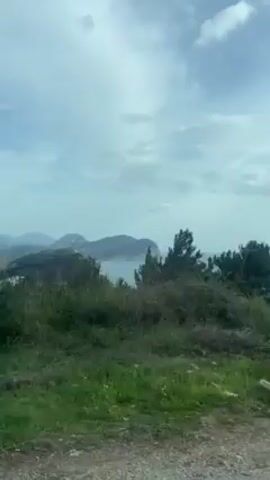The video shows a view from a moving vehicle, looking out towards a coastline under a cloudy sky. Green foliage lines the lower part of the frame, obscuring some of the view. In the distance, there are landmasses and a glimpse of the ocean. The focus is on the landscape and the overall scene.