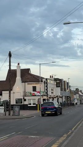 The video shows a street scene with a pub called "Crown & Anchor" on the left. A few cars are driving down the road, including a dark-colored Fiat 500 and a blue SUV. The sky is cloudy, suggesting overcast weather. The street appears to be in a suburban or town setting. There are utility poles and wires visible along the street, as well as streetlights. The pub has signage and some decorations visible. The video is filmed from a stationary point of view, looking down the road.