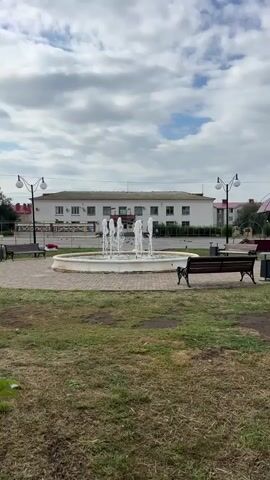 A white, circular fountain sprays water in the center of a paved area. Around the fountain are several dark-colored benches and lampposts with white globes. In the background, a long, white, two-story building with many windows is visible. The sky above is cloudy with patches of blue. The foreground consists of a grassy area.