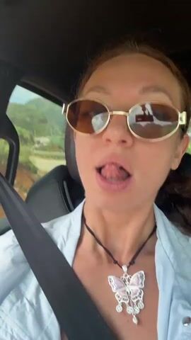 A woman with red hair and gold-framed sunglasses is in the driver’s seat of a car, wearing a light blue jacket, a black necklace with a butterfly pendant, and a seatbelt. The background shows a passing forest. Another person is visible in the backseat. The woman talks into the camera, then makes a surprised face. Music plays.
