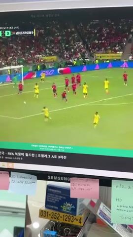 The video displays a computer monitor showing a live broadcast of a soccer match. The scoreboard indicates the game is between the Korean Republic and Ghana, with the score at 0-0. The players are wearing red and yellow jerseys. The camera is positioned above and behind the monitor, capturing the screen and the surrounding desk area. The desk is cluttered with various items, including sticky notes with handwritten text and drawings, a Samsung monitor, a metal container with office supplies, and what appears to be a phone or some sort of electronic device. The lighting suggests an indoor environment. The video's timestamp shows a duration of 5 seconds.