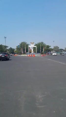 The video shows a wide, paved road with several cars driving on it. In the distance, there is a monument with an eagle symbol at the top and the words "TAMAN PANCASILA" written on it. There are trees and flags surrounding the monument. Orange traffic cones are placed on the road, creating a lane separation. The sky is clear and blue. The overall scene appears to be a public space, possibly a city square or an entrance to a park, on a sunny day.