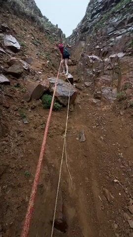 The video shows a person climbing up a steep, rocky slope using a rope. The slope is composed of loose soil, rocks, and small patches of vegetation. The person is wearing a red shirt, dark shorts, white sneakers, and a backpack. They are holding onto a thick, red and white rope that stretches down the slope. A thinner, white rope is also visible. The camera is positioned below the person, looking upwards, capturing their ascent. The camera then pans to the right, revealing a rugged cliff face. The camera continues to pan, showing a view of a beach below, with pebbles and sand, and the ocean in the distance. Several boats are on the beach. The sky is overcast.