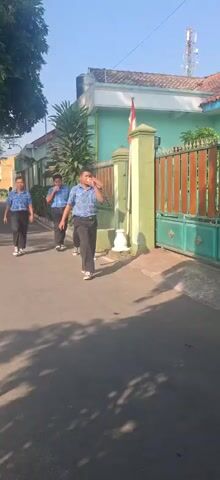 Four young males, appearing to be school-aged, are walking in single file on a paved path adjacent to a green fence. They are all wearing blue patterned shirts and dark pants. The male in the front right of the frame is seen wiping his face with his hand. The sun is casting shadows on the ground, indicating it is daytime. The environment appears to be an outdoor, possibly residential or school, setting with a building visible behind the fence.