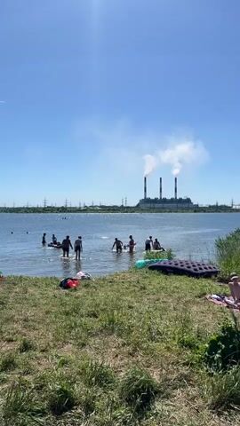 A group of people are seen swimming and relaxing on the shore of a lake. In the background, a large industrial facility with three smoking chimneys is visible under a clear blue sky. The shore is covered with green grass and scattered with beach towels and inflatable toys. Some individuals are wading in the shallow water, while others appear to be swimming further out. The overall scene suggests a warm, sunny day at a recreational area near an industrial site.
