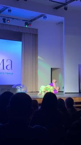 The video captures a dance performance on a stage viewed from the audience's perspective. The stage features two young girls performing a dance routine in matching neon green and purple outfits. One girl is lying on her back on the stage while the other is doing gymnastics poses while using the body of the other to support herself. In the background, there's a screen with text and a set of cream-colored curtains to the side. A silvery inflatable ball sits on the edge of the stage, illuminated by the stage lighting. The room is lit with blue and purple stage lights, creating a dramatic atmosphere. The audience is visible as silhouettes in the foreground.