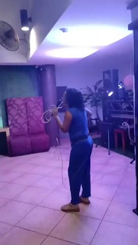 A woman in a blue jumpsuit stands on a tiled floor in a dimly lit room. She holds a microphone to her mouth, appearing to sing or speak. A coiled cable runs from the microphone to unseen equipment. The room has purple upholstered furniture and a ceiling fan. Colored lights illuminate the scene in alternating shades of pink, red, and green.