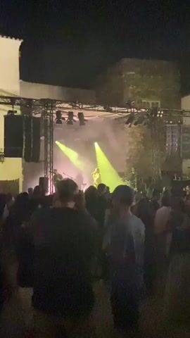 A nighttime outdoor concert is shown from behind a crowd of people. The stage is lit with yellow and pink lights and a fog machine is being used. A band is performing on stage. The crowd is facing the stage and some people are dancing. The background shows buildings and a dark sky.