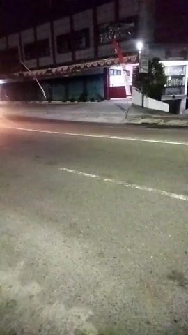 The video shows a street at night. The road is paved with asphalt and has a white line marking. On the left side of the road, there is a building with several windows and lights on. There are also some trees and streetlights visible. The right side of the road is bordered by a sidewalk and a fence. The overall impression is that of a quiet urban street at night.