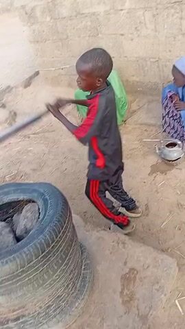 A young boy operates a water pump. The water pump is mounted on a pile of old tires. Three plastic buckets fill with water as he works the pump handle, standing on a stone.  Two young girls in hijabs watch nearby. One is dressed in a light green hijab and the other in a blue one. A small pot sits beside the girl in the blue hijab. The boy is wearing a black tracksuit with a red stripe. A brick wall forms a backdrop to the scene.