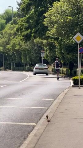 The video captures a sunny day scene on a road in Esch-sur-Alzette, Luxembourg. A person is riding an electric scooter on the right side of the road, wearing a backpack. A white car is ahead of them in the same lane, and another car, a dark grey SUV, is in the left lane, passing the scooter rider. The road is bordered by a sidewalk and lush green trees and hedges. Traffic signs are visible, including a yellow diamond-shaped sign and a red and blue circular sign. The time of day appears to be daytime, judging by the bright sunlight and shadows. The overall mood is peaceful and ordinary, depicting everyday life in the city.