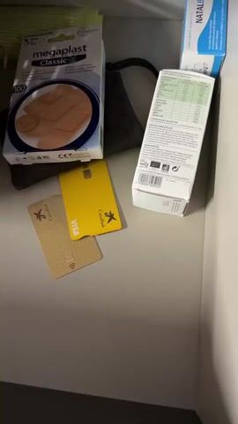 The video shows a shelf in a drawer or cabinet. On the shelf are several items including a box of bandages labeled "megaplast Classic", two credit cards, one yellow and one gold, both with the Visa logo. There is also a white box with nutritional information on it and a box labeled "NATALBEN". In a woven basket, there are several bottles and packages of what appear to be medications and toiletries, including blister packs of pills, white jars, and tubes of cream. A hand reaches into the basket and touches some of the items. The lighting is dim and the focus shifts throughout the video.