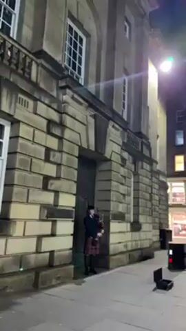 A man dressed in traditional Scottish attire is playing the bagpipes on a city street at night. The man is wearing a black jacket, kilt, and black boots. He is standing in front of a large stone building, which is illuminated by streetlights. There is a small black case next to him on the ground. The street appears empty of other people.