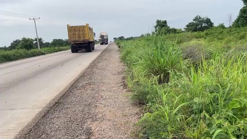 The video shows a road with a dirt shoulder and lush green vegetation on the right side. Several large trucks are traveling on the road, with one truck, a yellow dump truck, in the foreground. Further down the road, two more trucks are visible, one appears to be carrying a load of coiled wire. A motorcycle with a rider is also seen traveling on the left side of the road, passing the trucks. The sky is overcast, suggesting cloudy weather. The road appears to be in a rural or semi-rural setting, with trees and greenery visible in the distance.