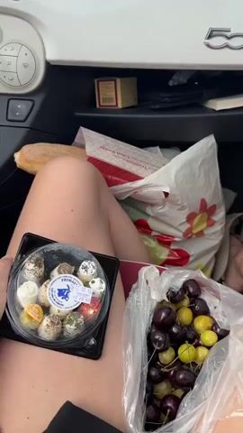 The video begins with a close-up view of a person's legs and lap inside a car. On the lap is a round plastic container filled with small, individually packaged cheeses, each appearing to have different toppings such as herbs or spices. Next to the cheese is a clear plastic bag filled with dark red and yellow cherries. In the background, a white paper bag with a red flower design is visible, along with other items in the car's glove compartment, including a box and a book. The car's interior, specifically the dashboard and a "FIAT" logo, is also visible.

The camera then pans to the windshield, showing the view outside the car. The car appears to be driving on a road surrounded by trees and greenery. There are road signs, one indicating directions to various towns, and a street lamp. A black car is seen in the distance, and a house with a distinct roofline is visible. The sky is partly cloudy, suggesting daytime. The video ends with the car continuing its journey on the road.