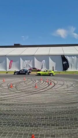 The video shows a bright, sunny day with a clear blue sky. The scene is an outdoor event, possibly a car show or competition, held on a paved area with interlocking brick patterns. Several cars are present on the pavement, including a black car with white lettering and a neon yellow car with racing modifications. A third car, possibly purple, is visible in the background. The cars are positioned around a circular track marked with orange cones. Tire marks are visible on the pavement, indicating that the cars have been drifting or performing stunts. A large, white tent structure is situated behind the track. In the distance, there is a crowd of spectators, some trees, and a line of vehicles. The overall atmosphere suggests an active and energetic event.