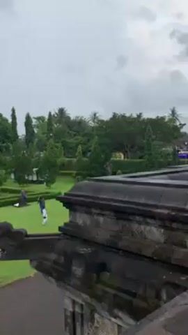 Here is a detailed description of the video:

The video shows a view from atop a stone structure, likely an ancient temple or monument, on a cloudy day. The camera slowly pans from left to right, revealing the structure's layered stone construction, intricately carved with decorative elements. In the distance, a well-maintained green lawn with manicured bushes and various trees can be seen. Several people are walking on the lawn, and a few buildings, including a structure with a red and blue awning, are visible further in the background. The overall atmosphere is subdued due to the overcast sky.