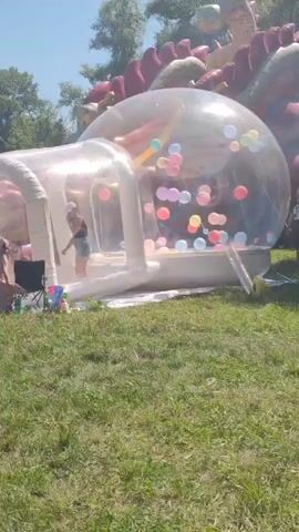 A group of people are gathered in a park on a sunny day. In the foreground, a large inflatable structure resembling a dragon is set up on a grassy field. The dragon has a large, clear, inflatable dome attached to it, filled with colorful balloons. Several people are visible around the inflatable structure, including a person wearing a dark uniform standing to the right of the dome and another person walking away from the camera in the center-left. In the background, trees and a clear blue sky are visible. The overall atmosphere appears to be festive and recreational.